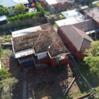 Aerial view of a partially demolished red-brick house with exposed roof beams and debris surrounding the property, situated between two intact houses—a stark reminder of ongoing home demolitions in the area.