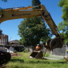 A large yellow excavator with the branding "Home Demolitions" is seen in a grassy yard, operated by a person in an orange vest and helmet. Debris and metal fencing are visible in the background.