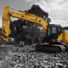 A yellow excavator with "Home Demolitions" on the arm is parked on a pile of rubble at a construction site surrounded by trees and buildings.