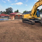 A yellow excavator is parked on a freshly cleared plot of land in front of residential houses, likely after recent home demolitions. The ground appears to be leveled and prepared for construction.